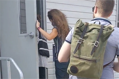 [ai] A person holding a door open for another individual as they enter a building. The second person has long, curly hair and is wearing a black and white outfit. A third individual with a backpack is standing nearby.
